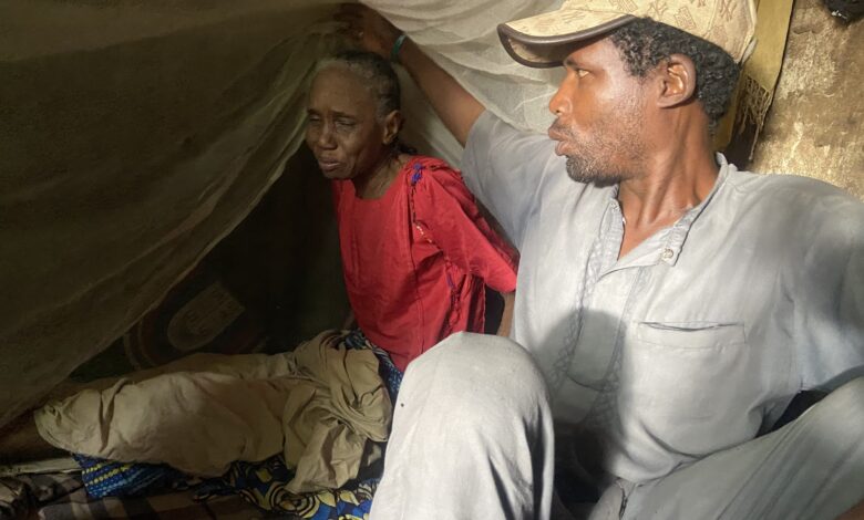 Two people sitting under a makeshift tent, one looking weary and the other gazing into the distance.