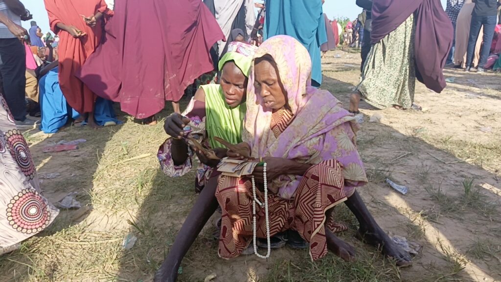 Two women in traditional attire examining money closely on a sunny day outdoors with other people in the background.