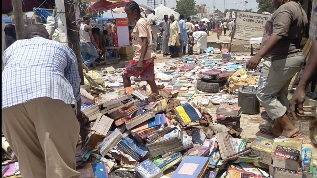 Bustling outdoor market scene with people browsing through piles of used books on the ground.