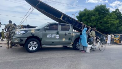 Army vehicle and civilians on a busy street with an overturned billboard in the background.