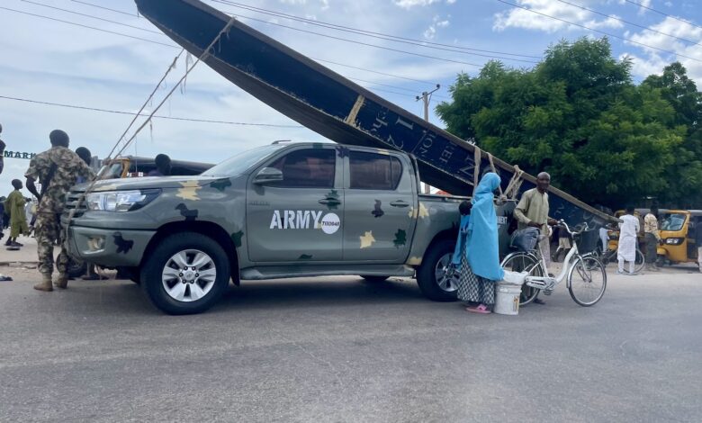 Army vehicle and civilians on a busy street with an overturned billboard in the background.