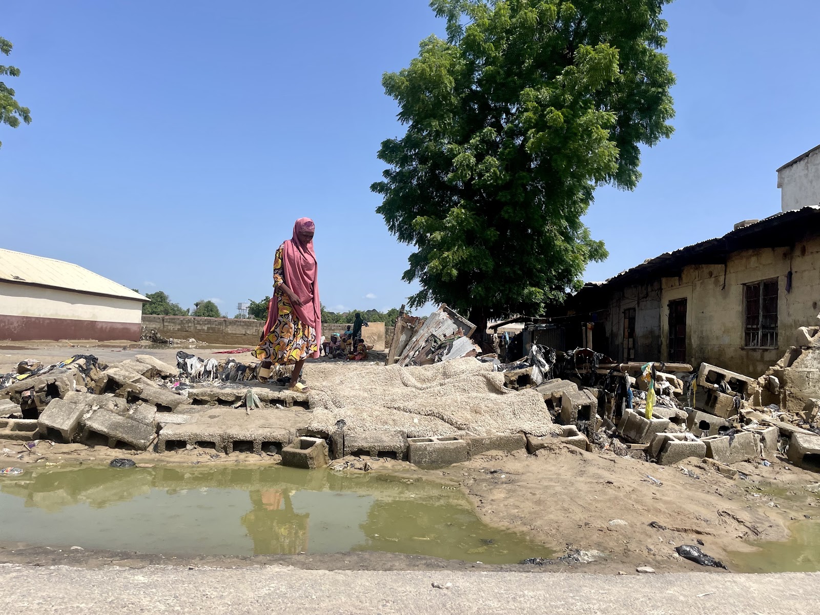 Person walking among rubble and standing water in a devastated area with ruined buildings and a tree in the background.