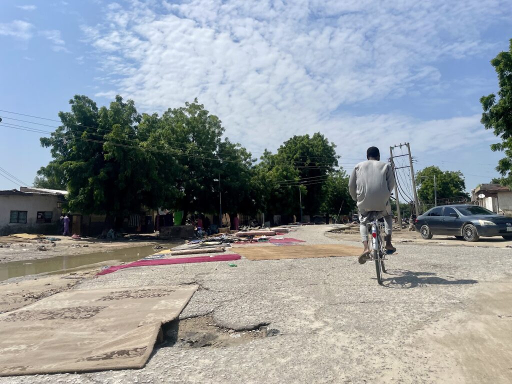 Person riding a bicycle on a damaged road with rugs laid out to dry and trees in the background under a cloudy sky.