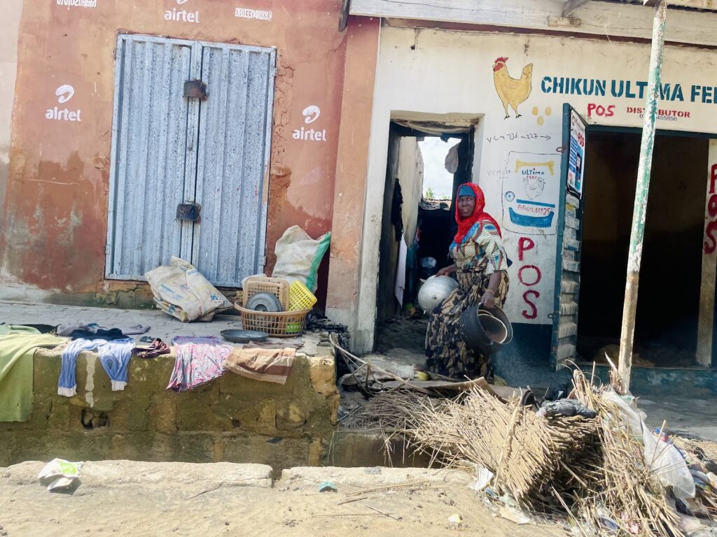 Woman standing by a doorway near a closed shop with advertisements, laundry drying, and debris on the ground.