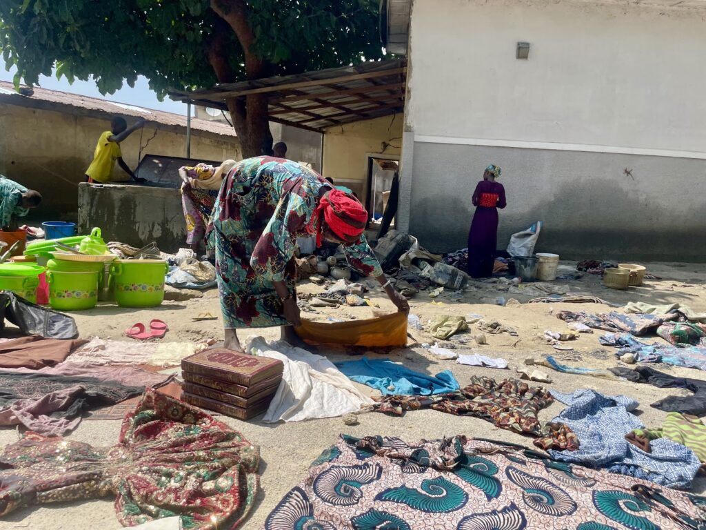 People sorting fabrics and items in a sandy outdoor area under a tree.