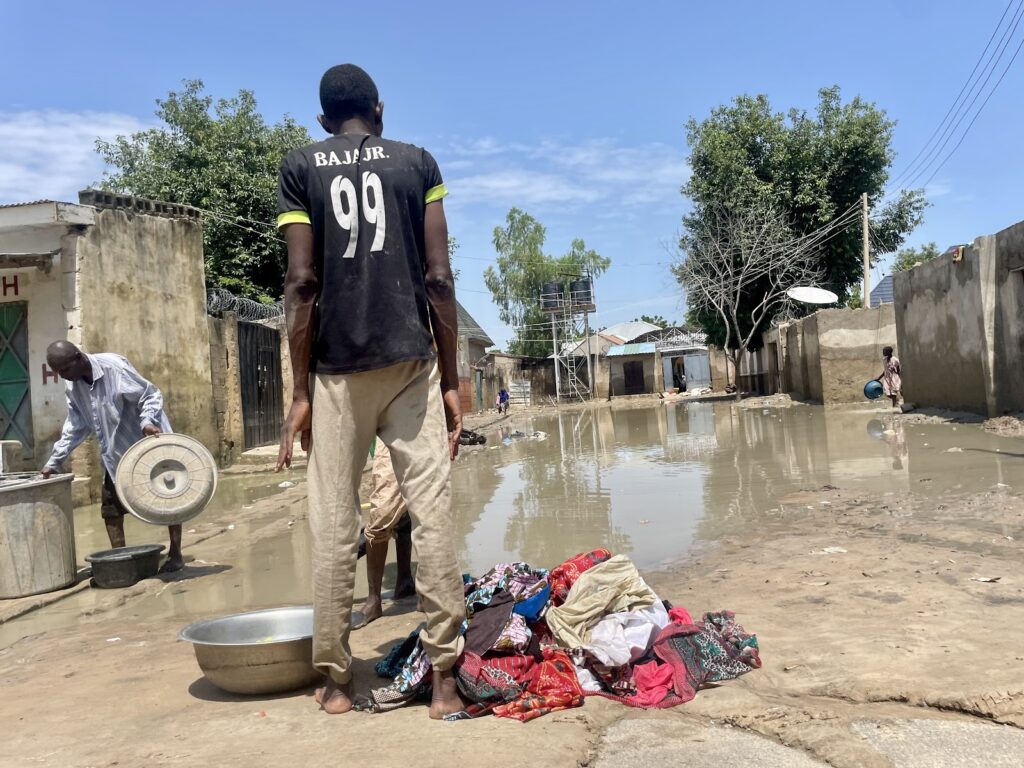 Individual standing in a flooded street with scattered clothes, while another person washes utensils nearby.