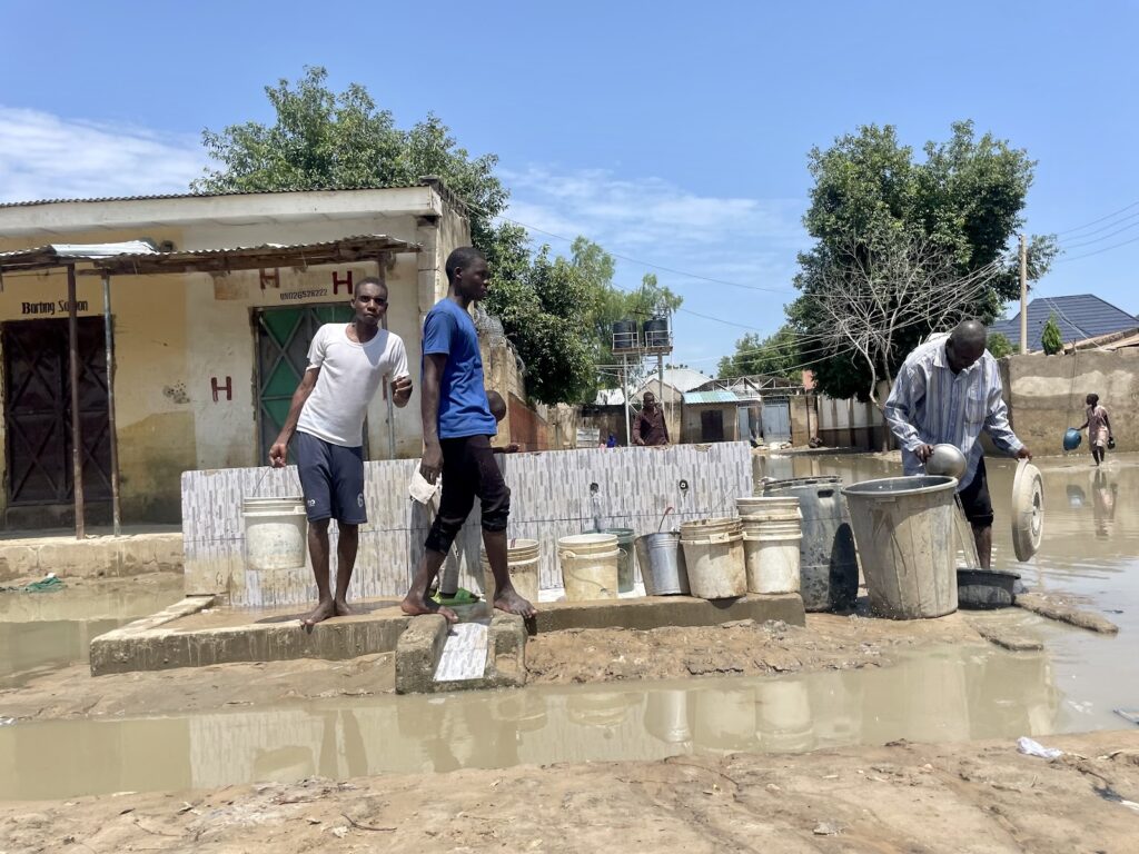 Two men navigating a flooded street with stepping stones while a third collects water in containers.