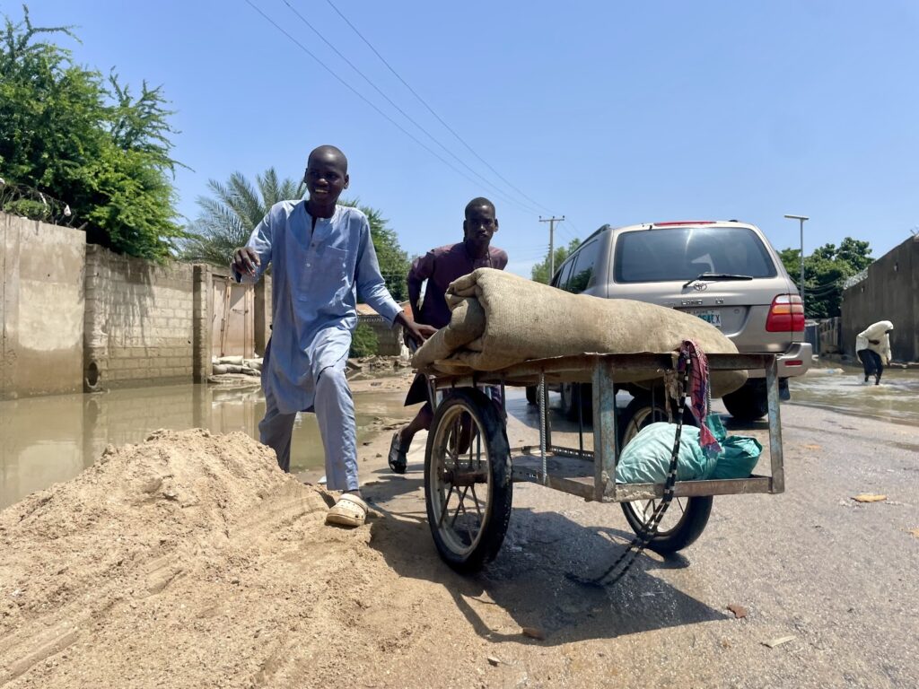 Two men with a loaded cart on a sunny street with puddles, a parked car, and a sandy mound in the foreground.