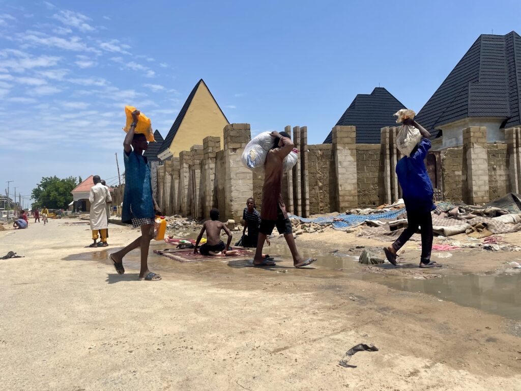 People walking with items on their heads near waterlogged area with buildings in the background.