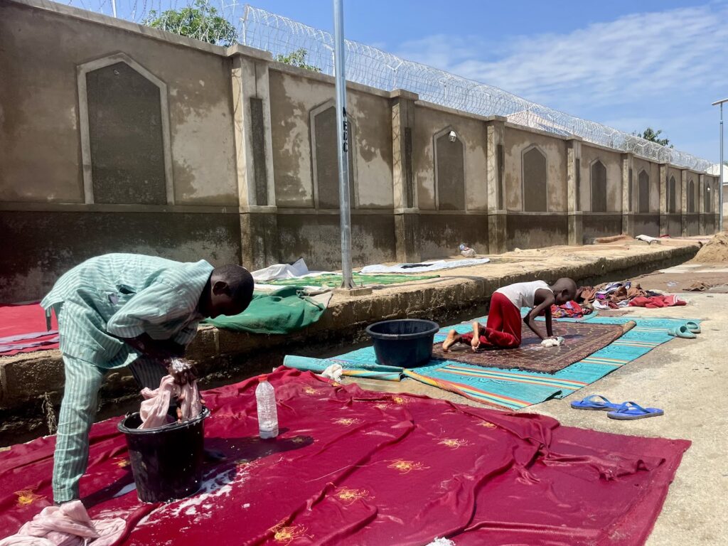 People washing clothes by hand on fabric spread outside under a bright sky, with a concrete wall and barbed wire in the background.