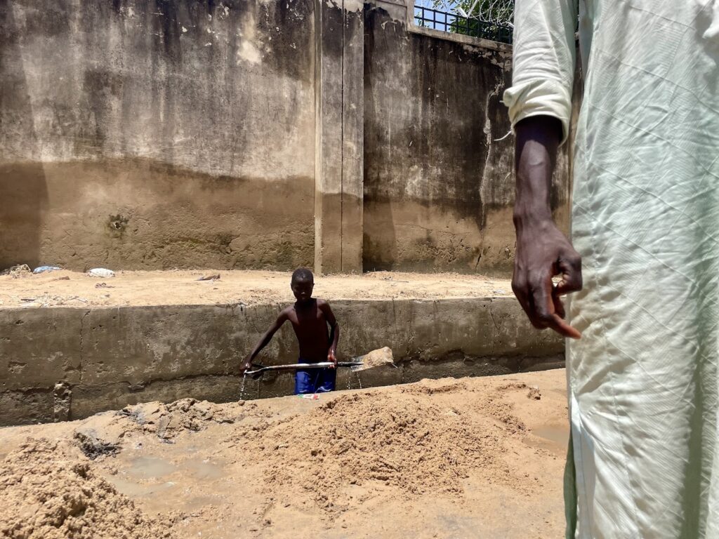 A child working in a sandy area by a wall, with the arm of an adult in the foreground.