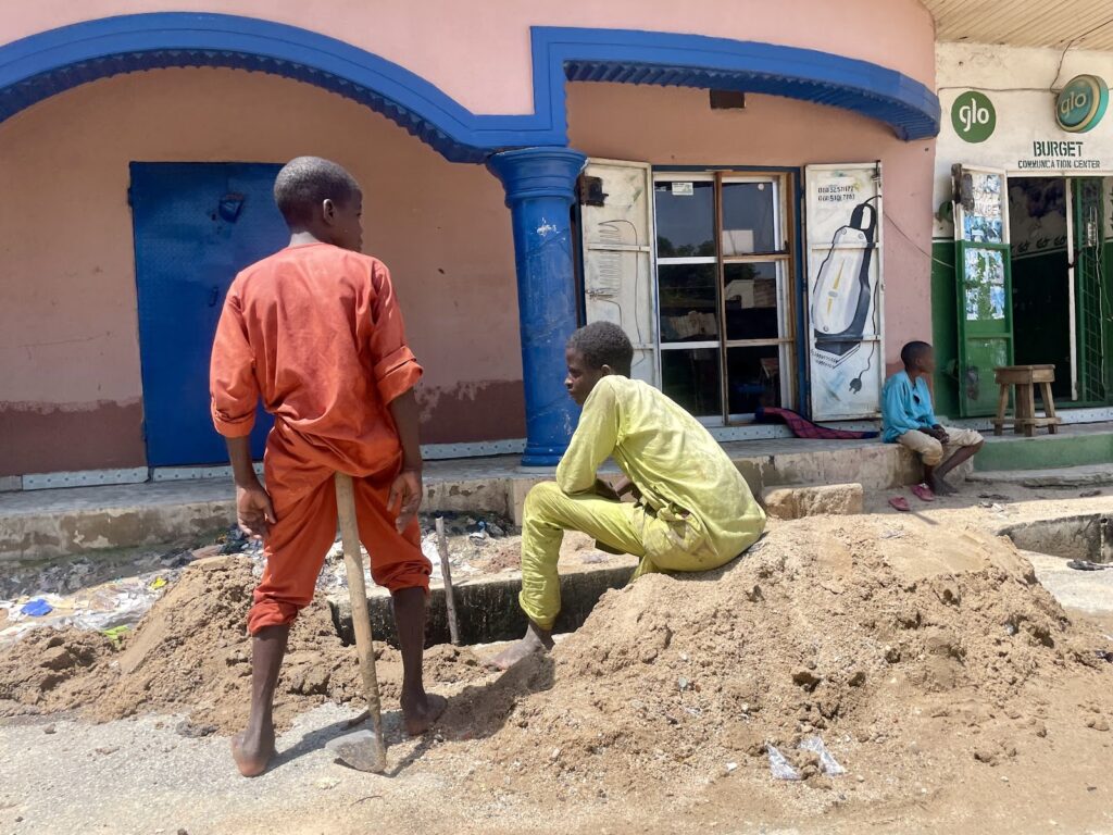 Two boys near a mound of dirt on a sunny day, one standing with a shovel, another sitting with buildings in the background.