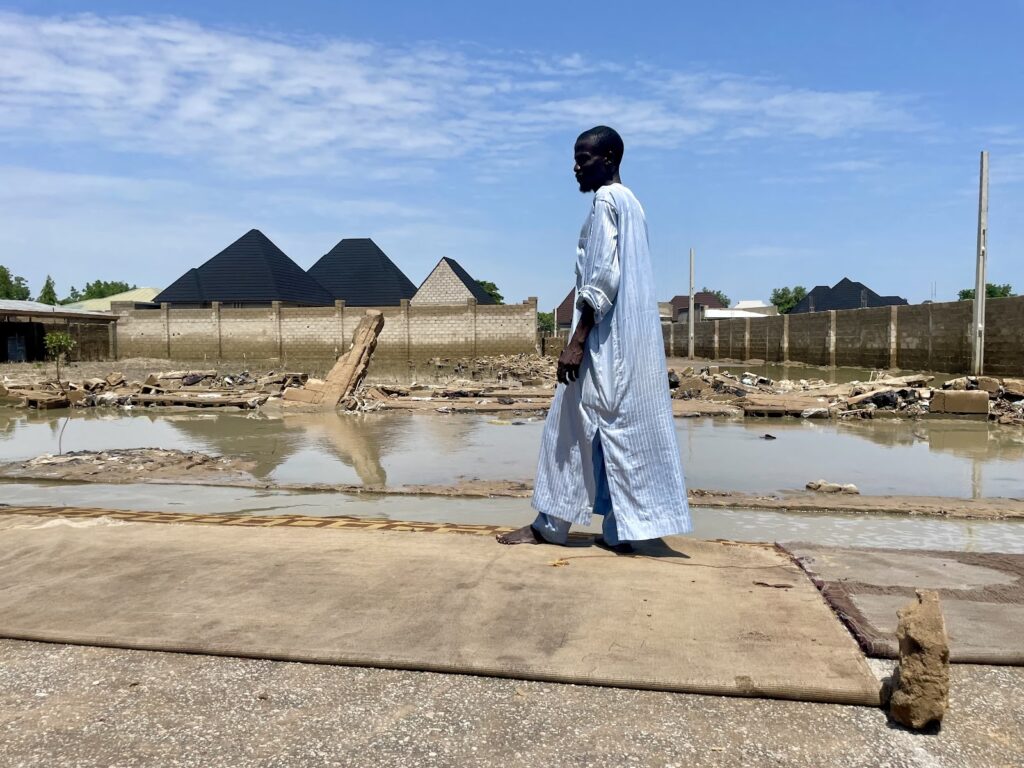 Person in blue garb walking by a flooded area with debris and buildings in the background.