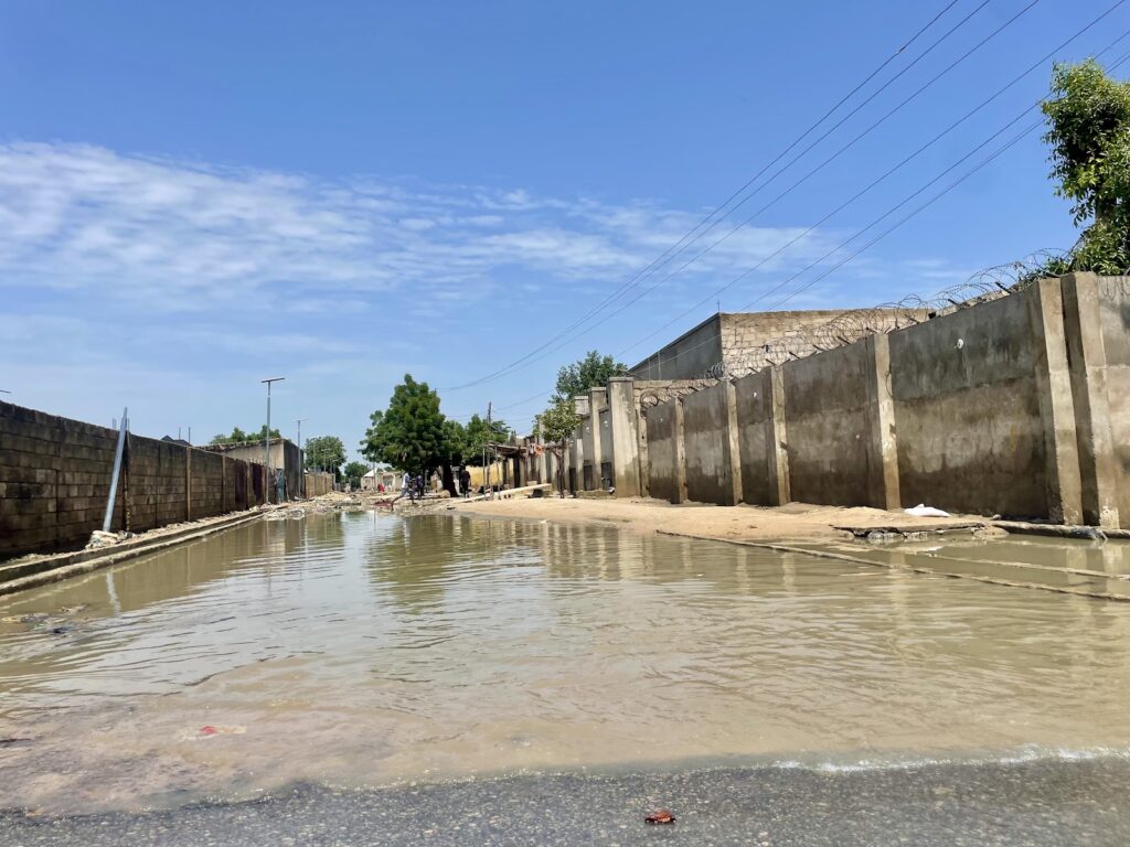 A flooded street with concrete walls under a blue sky, power lines above, and a few people in the distance.