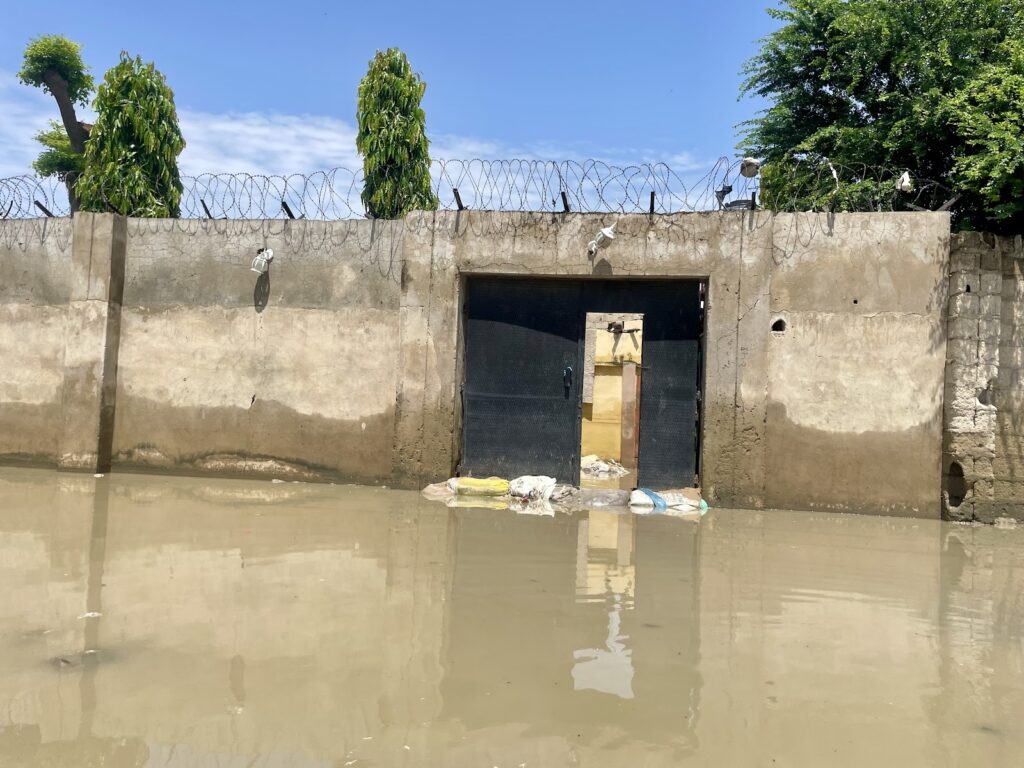 A flooded street with a high water level reaching a barricaded gate, CCTV cameras, and barbed wire on the wall.