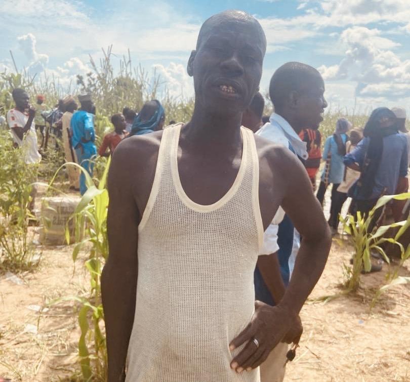 Man in white tank top standing in a field with other people in the background under a sunny sky.