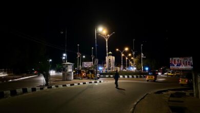 A person crossing a poorly lit street at night with vehicles and streetlights in the background.