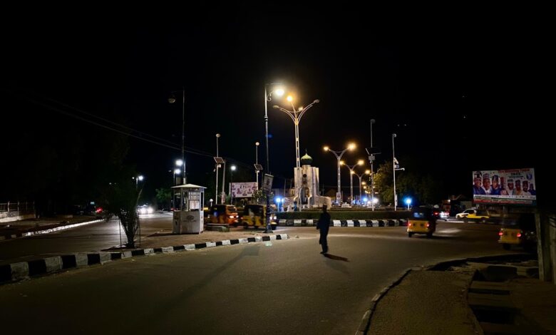 A person crossing a poorly lit street at night with vehicles and streetlights in the background.