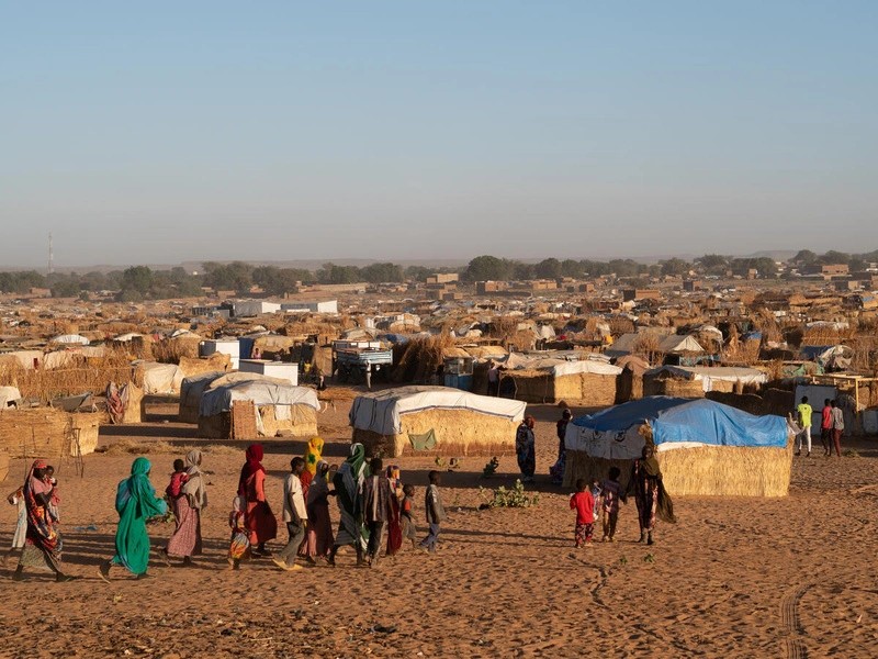 A bustling settlement with thatched huts and people milling about during golden hour.