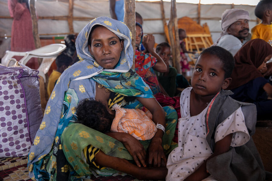 A woman nursing a child with another young child beside her, in a crowded tent setting.