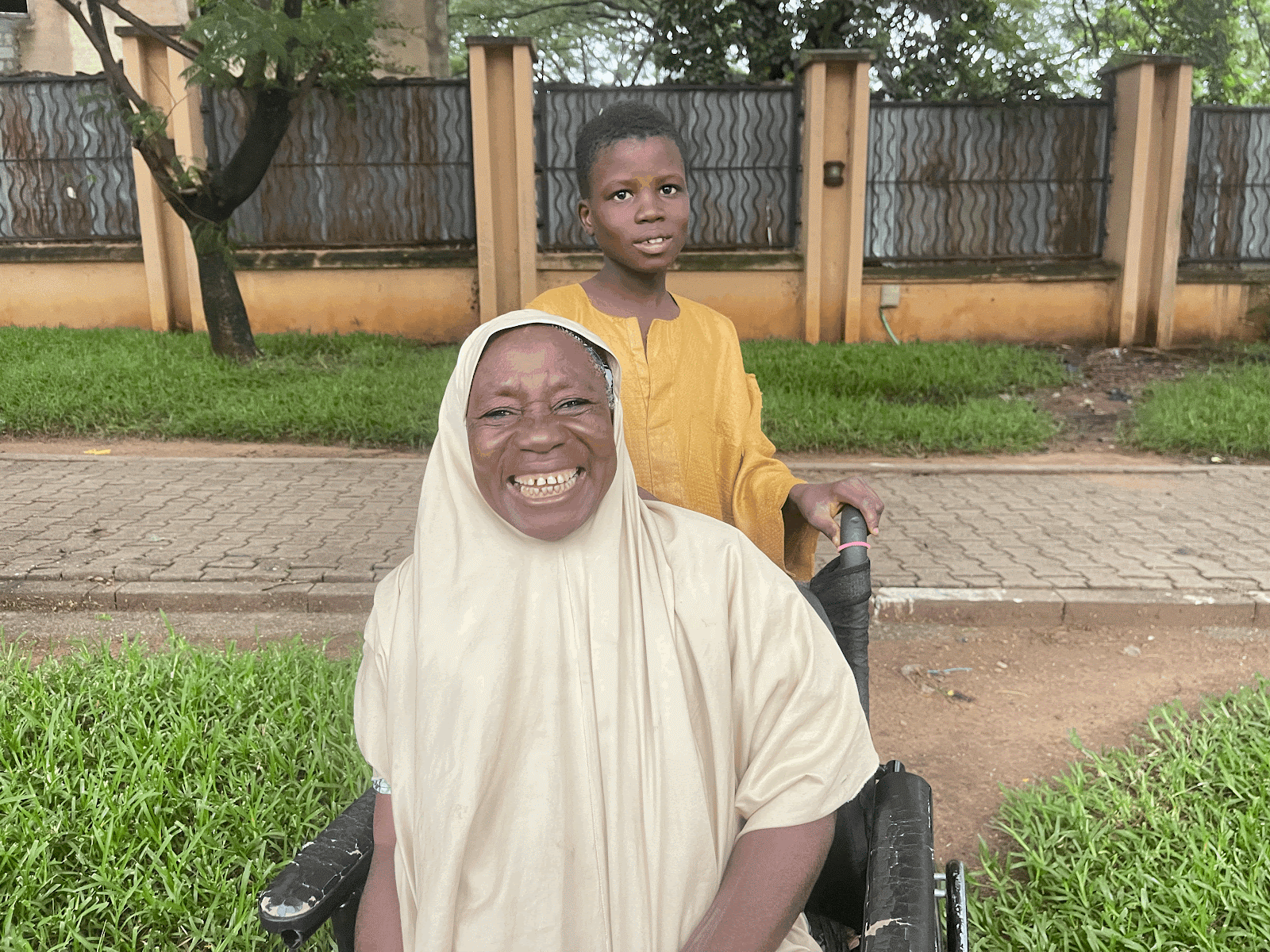 A joyful woman in a headscarf with a smiling child standing behind her, outdoors near a fence.