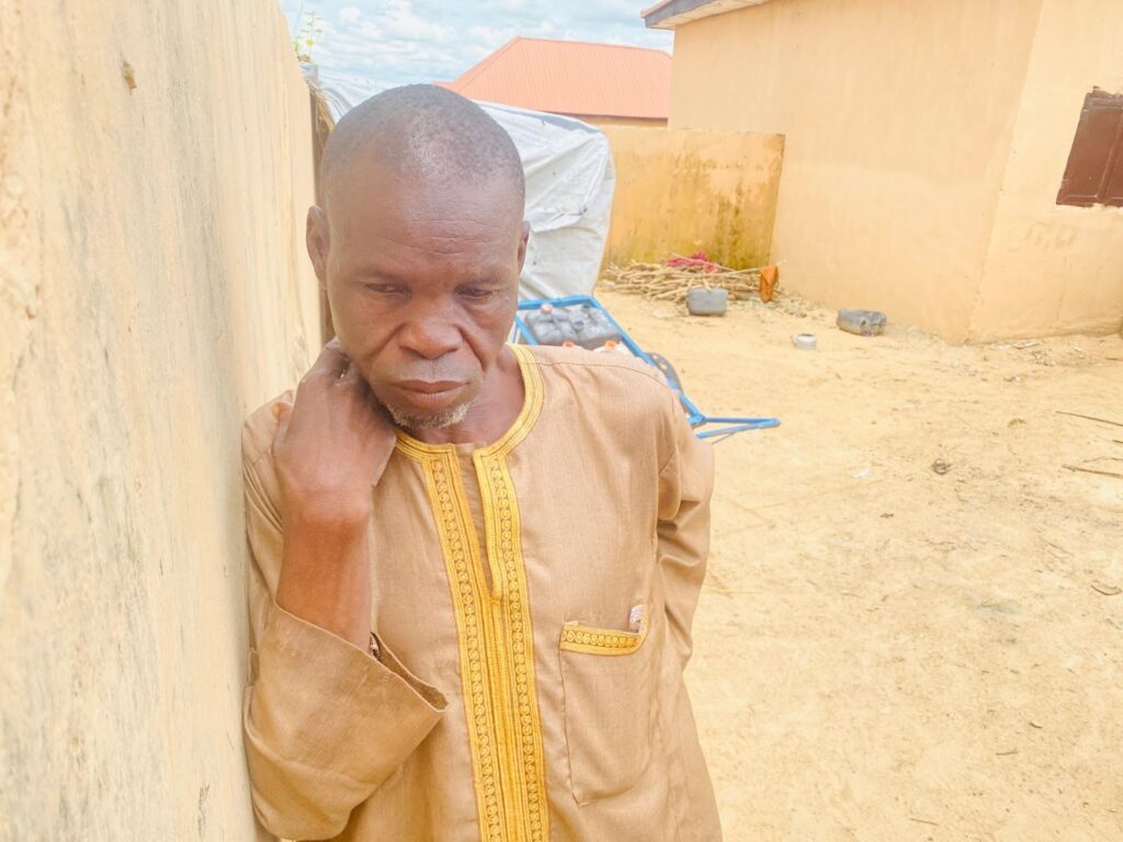 A man in a traditional outfit leaning against a yellow wall, looking thoughtful.