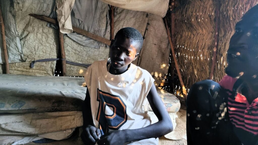 A young person sitting inside a traditional hut with dappled sunlight filtering through the thatch roof.