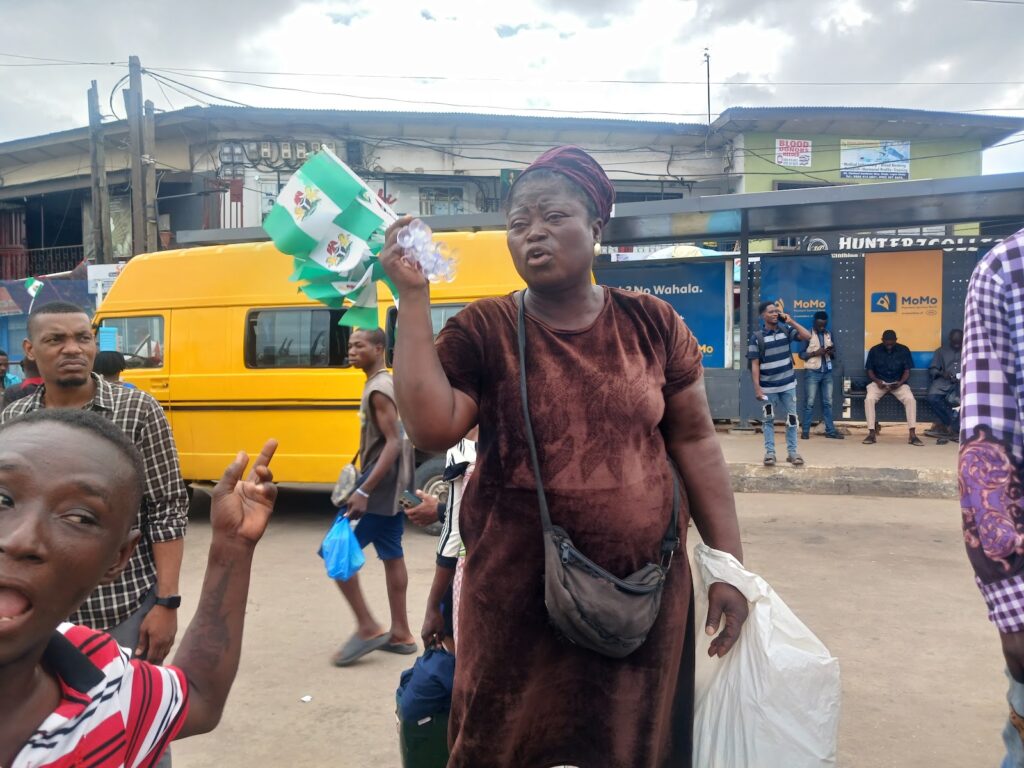 A woman holding up a plastic item on a bustling street with onlookers and a yellow bus in the background.