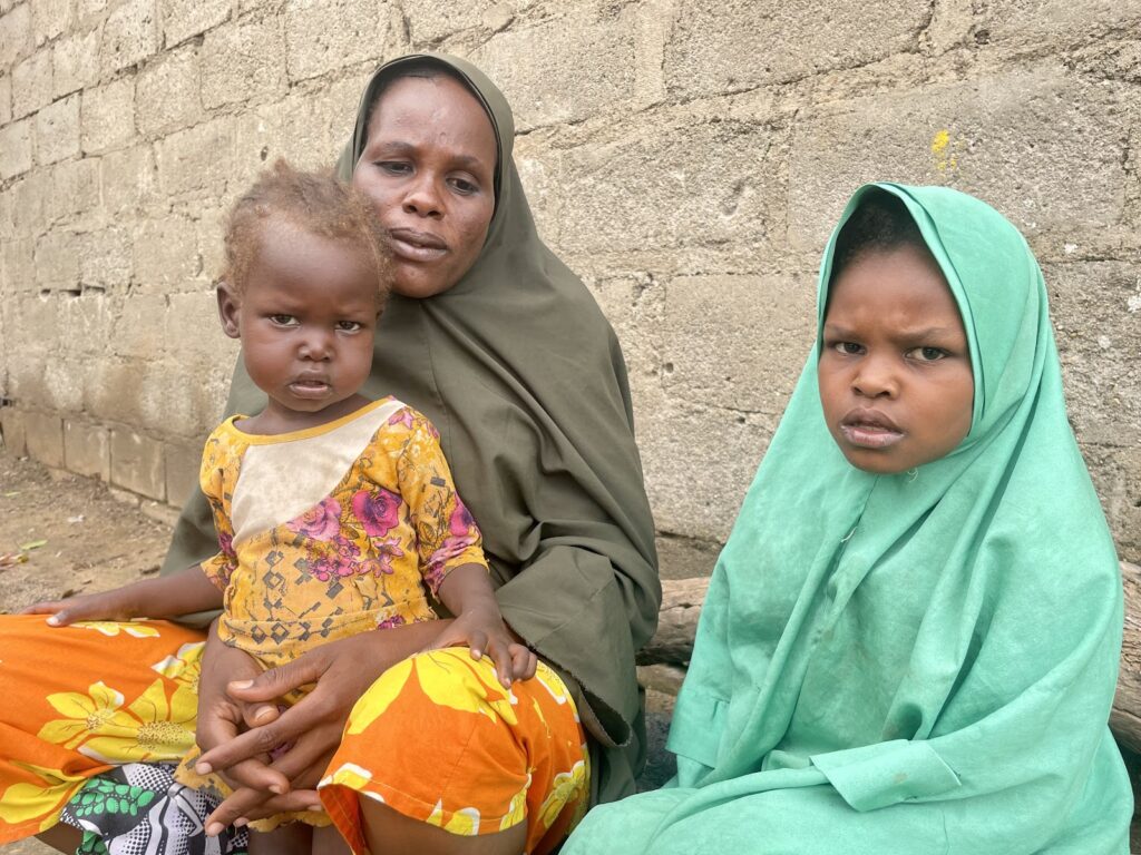 A woman with two children sitting by a wall, one child on her lap, the other beside her.