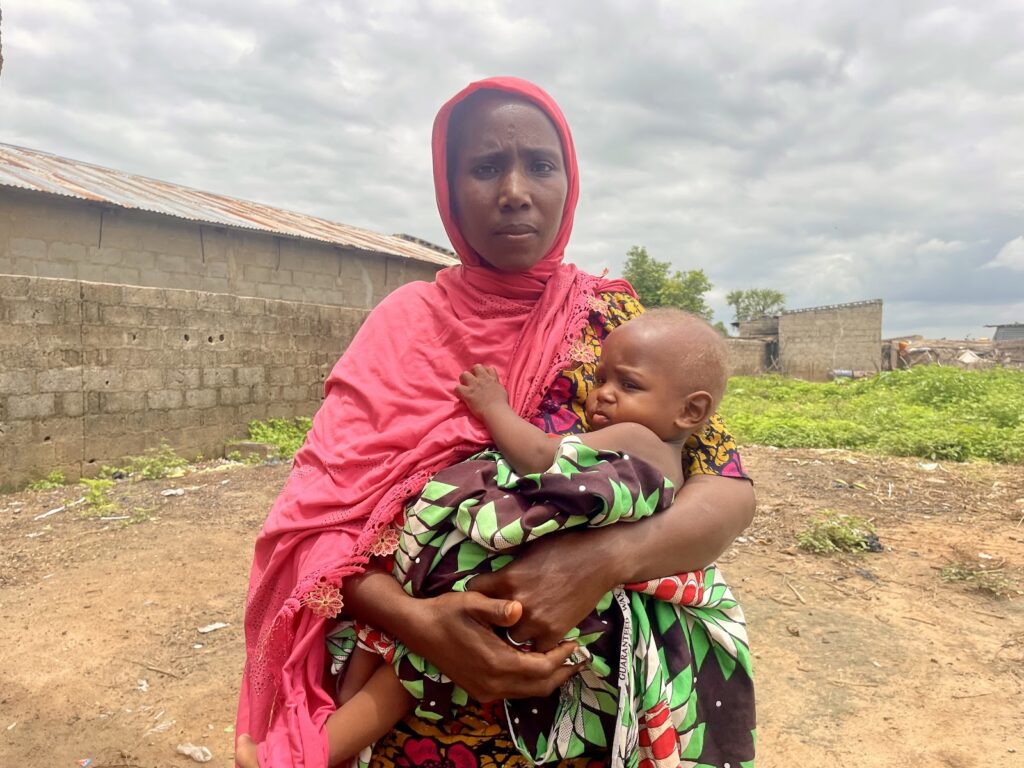 Woman in pink headscarf holding a sleepy child with a rural backdrop.