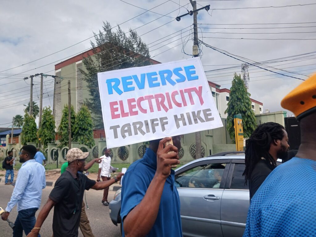 People marching with a sign reading "REVERSE ELECTRICITY TARIFF HIKE" on a street.