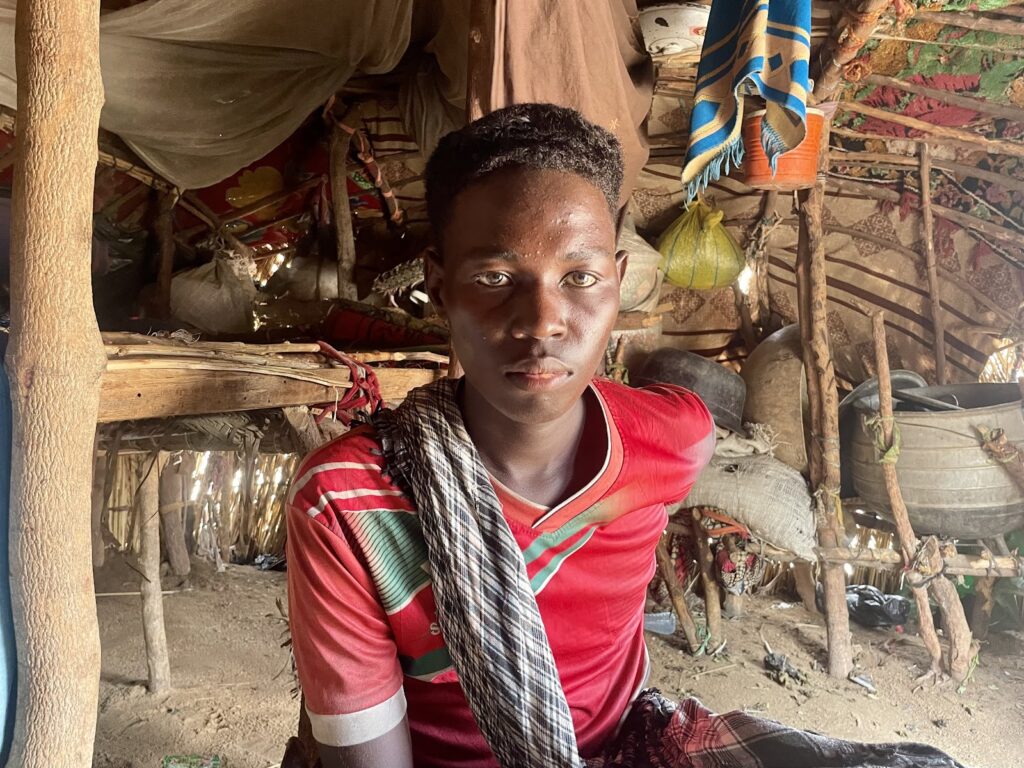 Young person in red shirt sitting in a rustic, traditional dwelling with various household items.