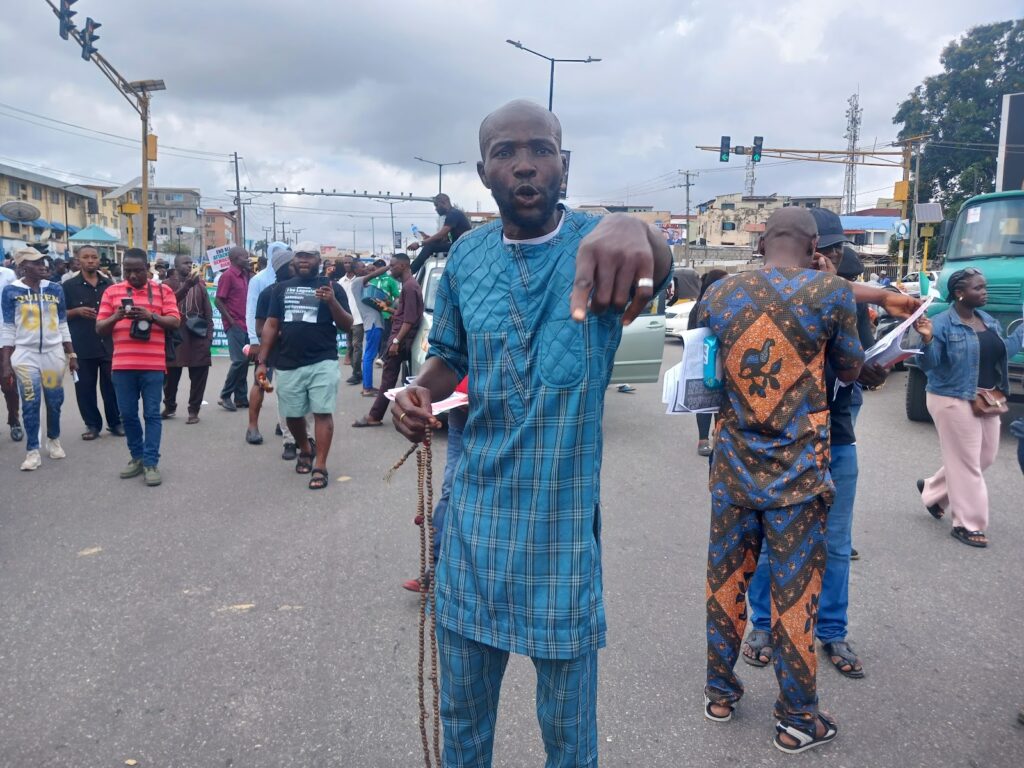 Man in blue traditional attire gesturing at the camera on a busy street with people.