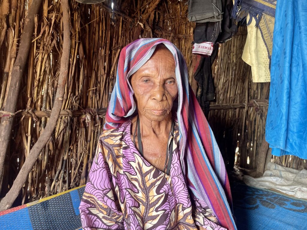 An elderly woman with a headscarf sitting inside a hut with dried reeds walls.