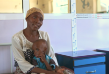 Woman in a headscarf sitting with small child on her lap, indoors near hospital beds.