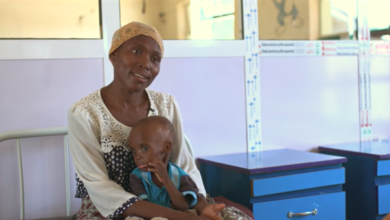 Woman in a headscarf sitting with small child on her lap, indoors near hospital beds.