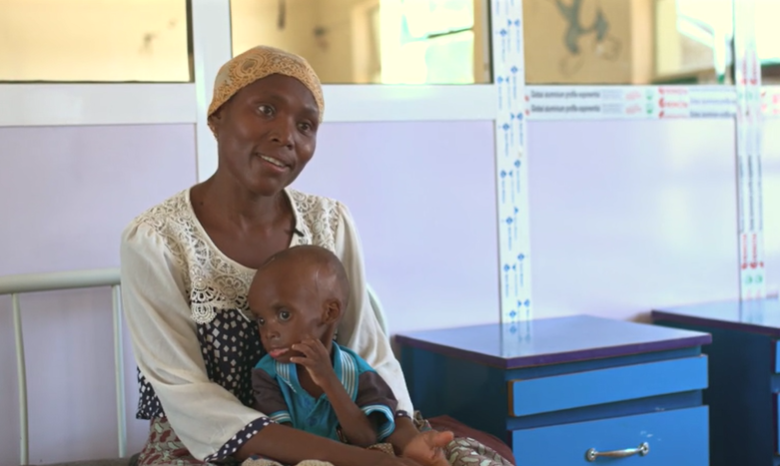 Woman in a headscarf sitting with small child on her lap, indoors near hospital beds.