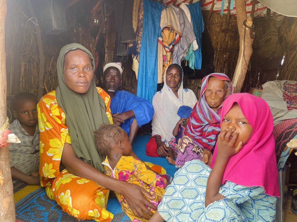 A family sitting together inside a hut with colorful clothing and expressions of mixed emotions.