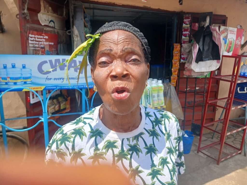 Elderly woman wearing a palm-print shirt talks in front of a shop with household items.