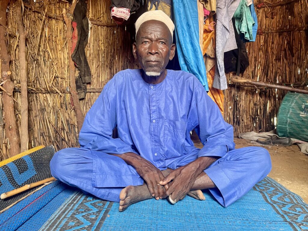 An elderly man in traditional blue attire sitting cross-legged inside a hut with thatched walls and clothes hanging in the background.