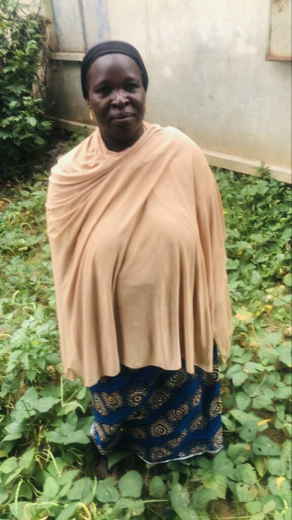 Woman standing in a garden wearing a beige shawl and blue patterned skirt.