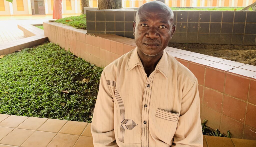 An elderly man wearing a striped shirt sitting in an outdoor area with a pink tiled wall and greenery.