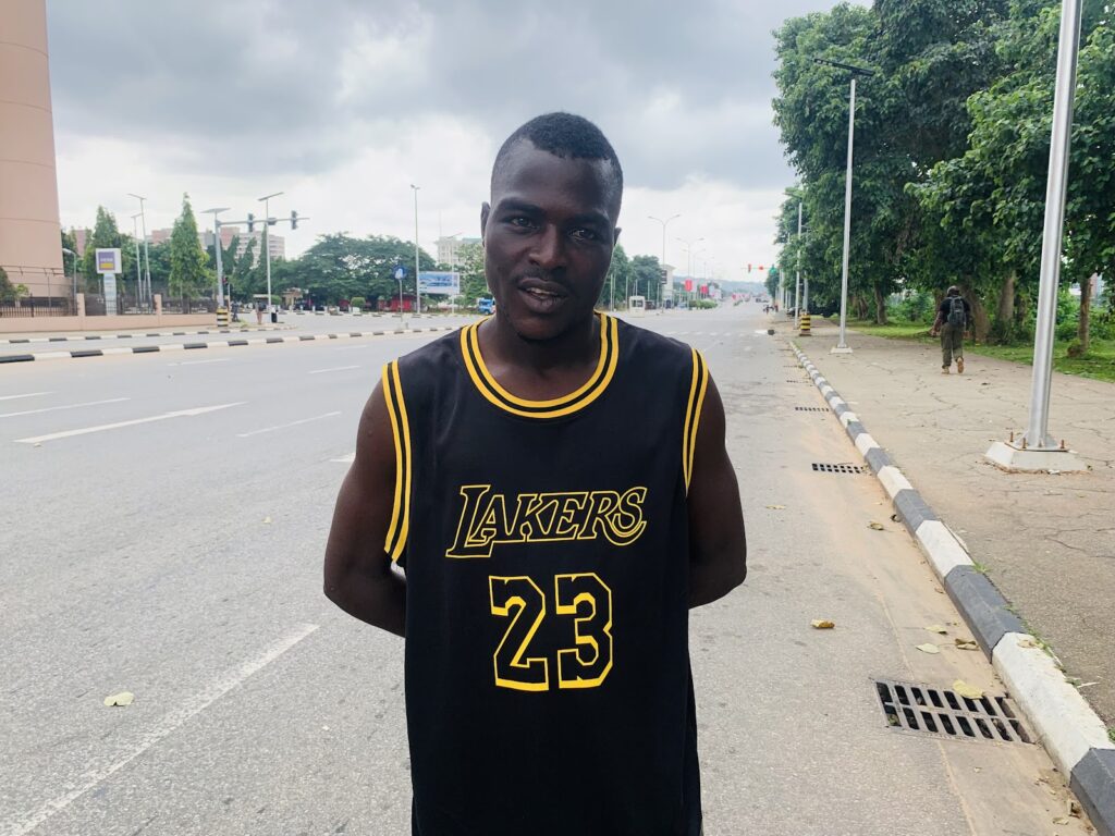 Man in a black Lakers jersey standing on an empty street with trees and traffic lights in the background.