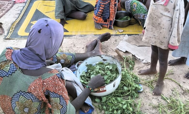 Person in a purple headscarf preparing greens with a child standing nearby on sandy ground.