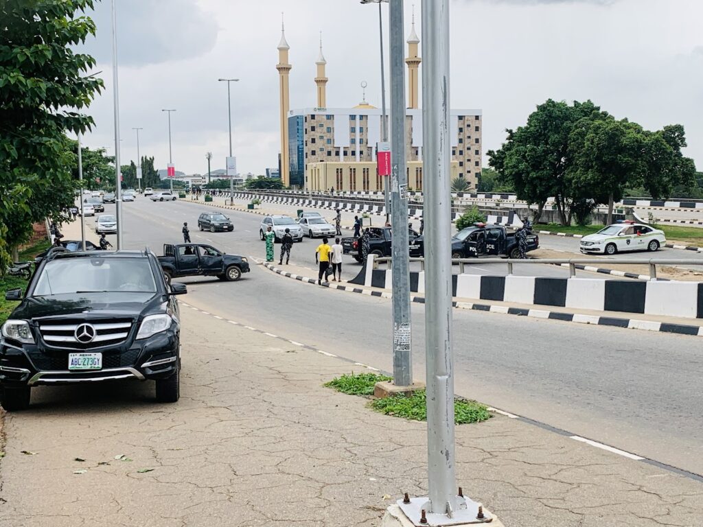 A busy street scene with cars, people walking, and a police presence near a median strip, with a mosque in the background.