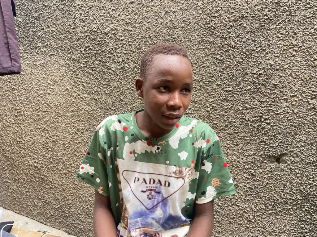 Young person in a green printed shirt standing against a textured wall.