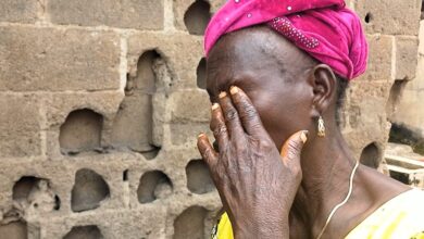 Woman with pink headscarf holding her face, showing emotion, against a cinder block wall.