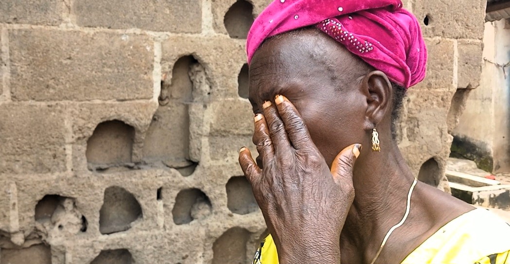 Woman with pink headscarf holding her face, showing emotion, against a cinder block wall.