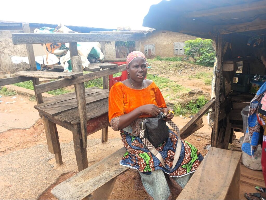 Woman in an orange blouse and patterned skirt seated on a bench outside a rustic stall, holding a cloth.