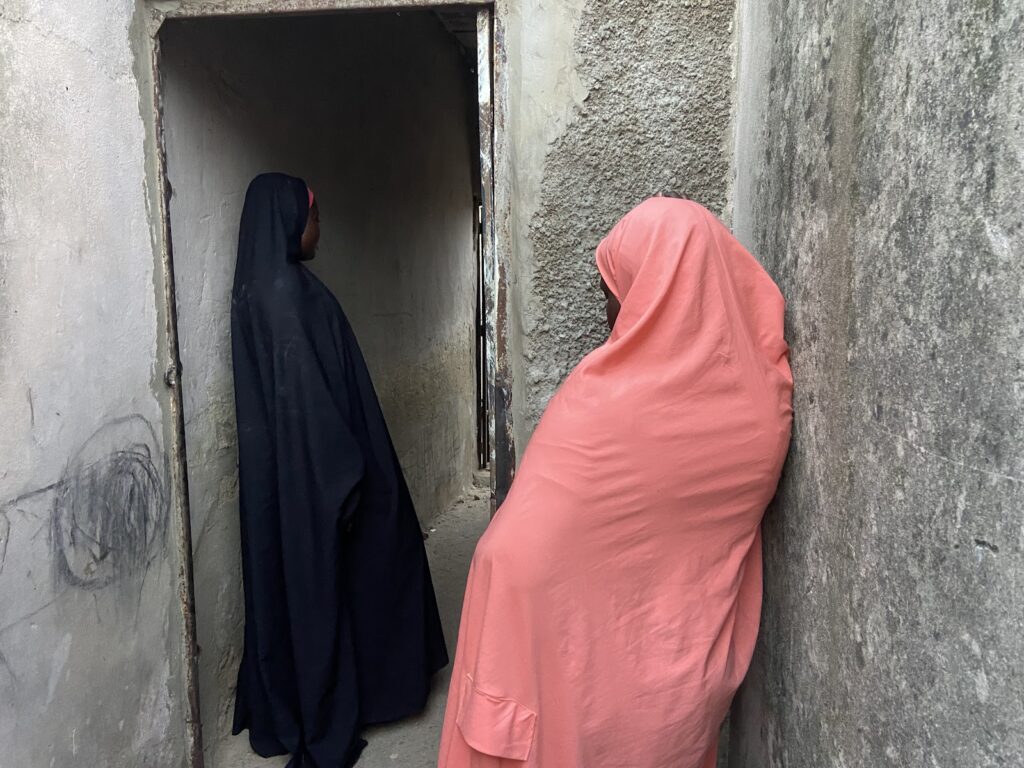 Two people in traditional attire standing by a doorway and a wall in a narrow alleyway.